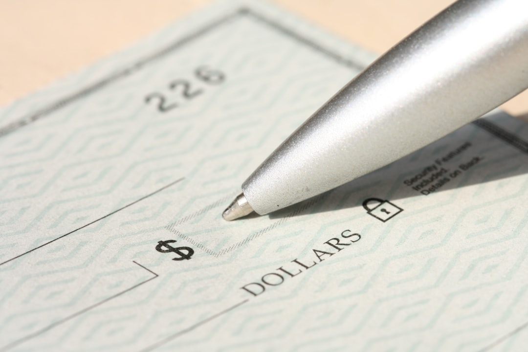 A close-up, high-resolution image of a blank check with a security pattern, showing the dollar sign box and part of the “DOLLARS” line. A metallic ballpoint pen rests near the writing area.
